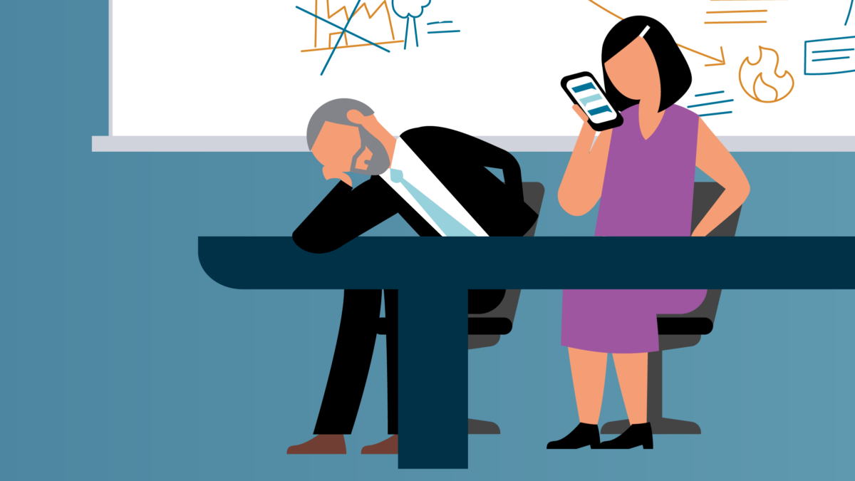 Two individuals sit at a table in front of a busy whiteboard. One stares at their phone, the other is slumped over and asleep.