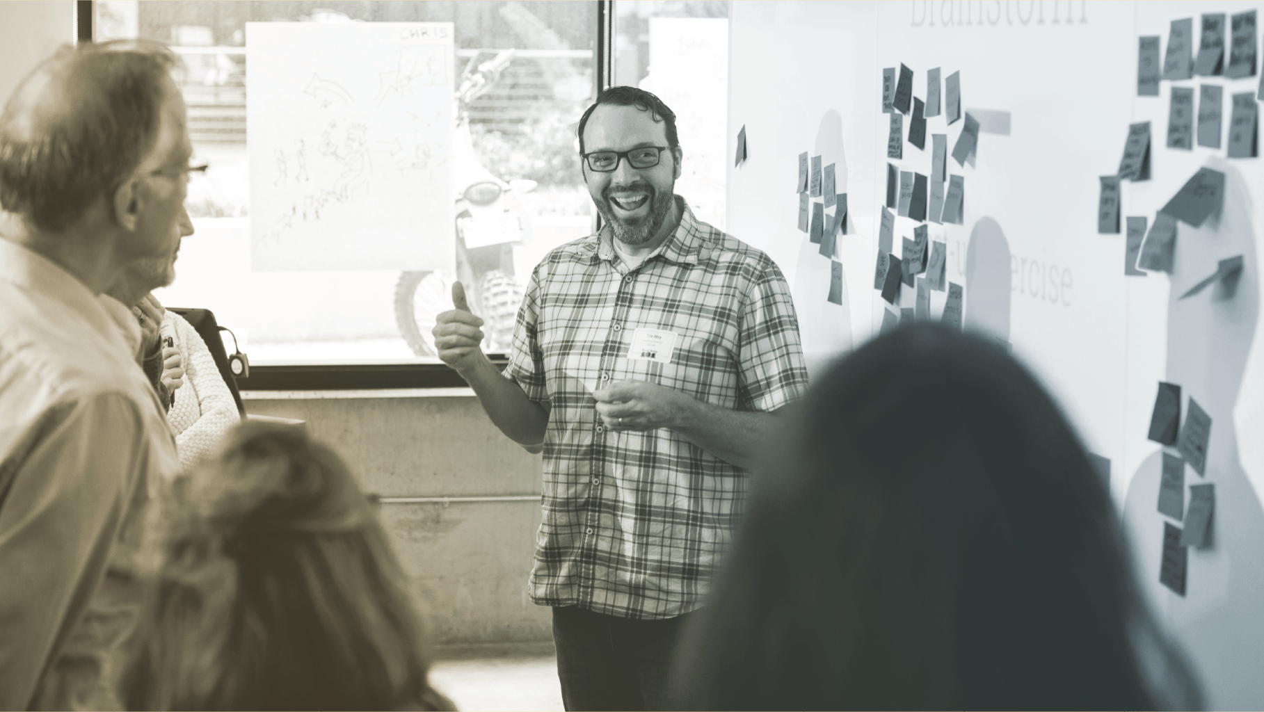 Smiling man with a thumbs up stands in front of several others, all in front of a whiteboard with sticky notes covering it