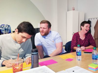 A group of three individuals sit around a table with lots of sticky notes and water bottles, laughing and discussing